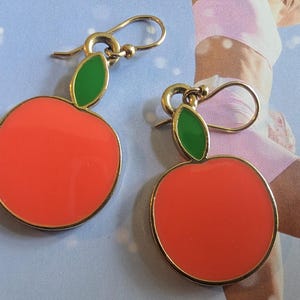 May include: A pair of apple-shaped earrings. Each earring features a bright red enamel apple with a gold-colored border and a green leaf at the top. The earrings have gold-colored ear wires.
