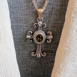 May include: A silver-toned cross pendant necklace with a gold-toned chain. The cross features an oval-shaped, dark-coloured stone surrounded by small, clear stones. The cross has an ornate, decorative design.