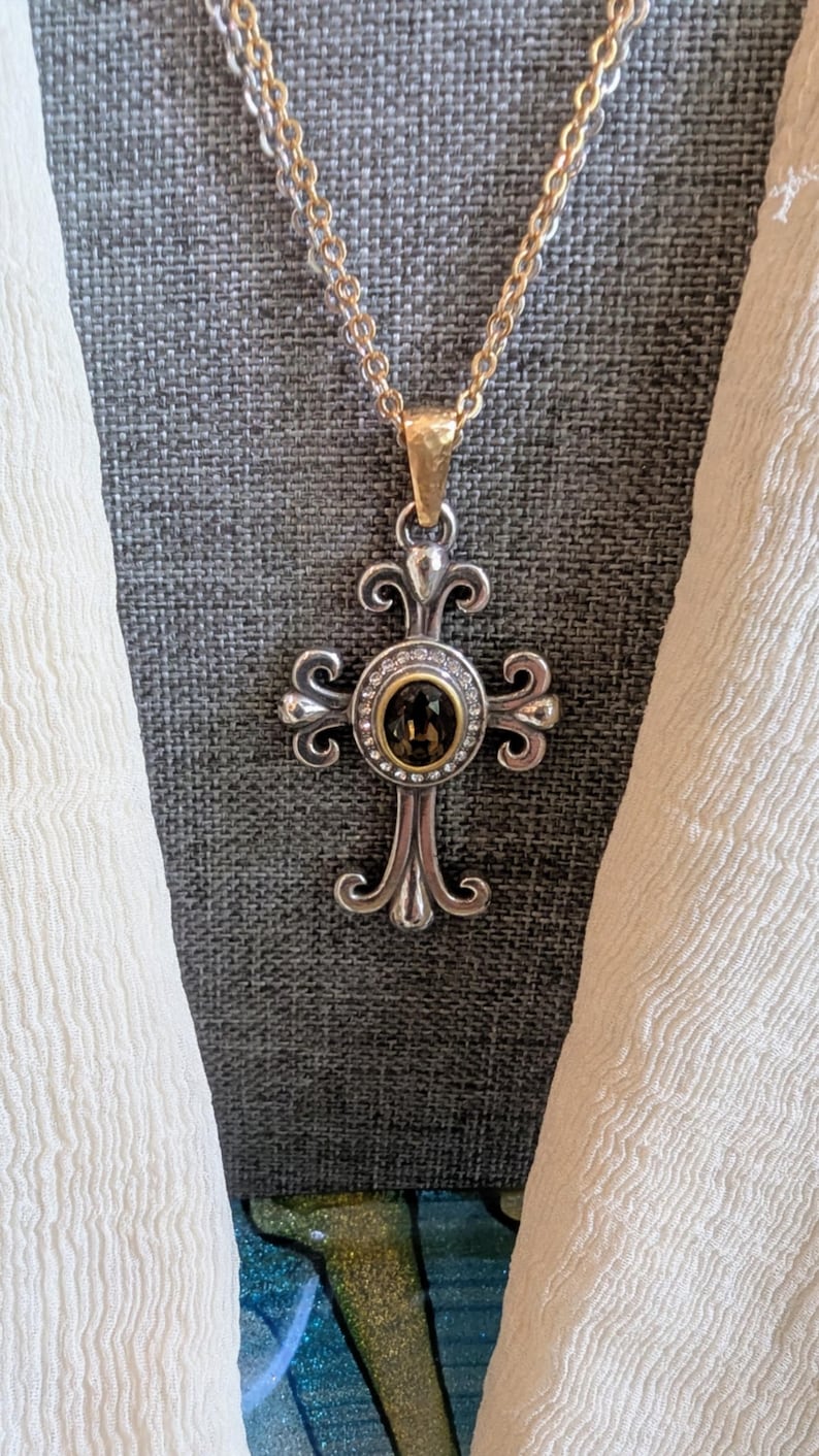 May include: A silver-toned cross pendant necklace with a gold-toned chain. The cross features an oval-shaped, dark-coloured stone surrounded by small, clear stones. The cross has an ornate, decorative design.