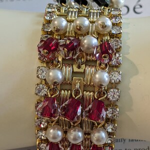 May include: A gold-toned bracelet with alternating rows of clear rhinestones, faux pearls, and faceted red crystals. The bracelet also features black crystal accents and a woven design. A vintage piece of jewelry.