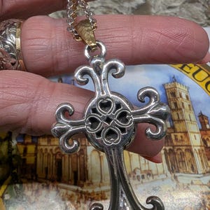 May include: Silver-coloured cross pendant necklace with a decorative design. The cross has a detailed, openwork centre with heart-shaped cutouts and ornate, curved arms. The chain is a mix of silver and gold tones.