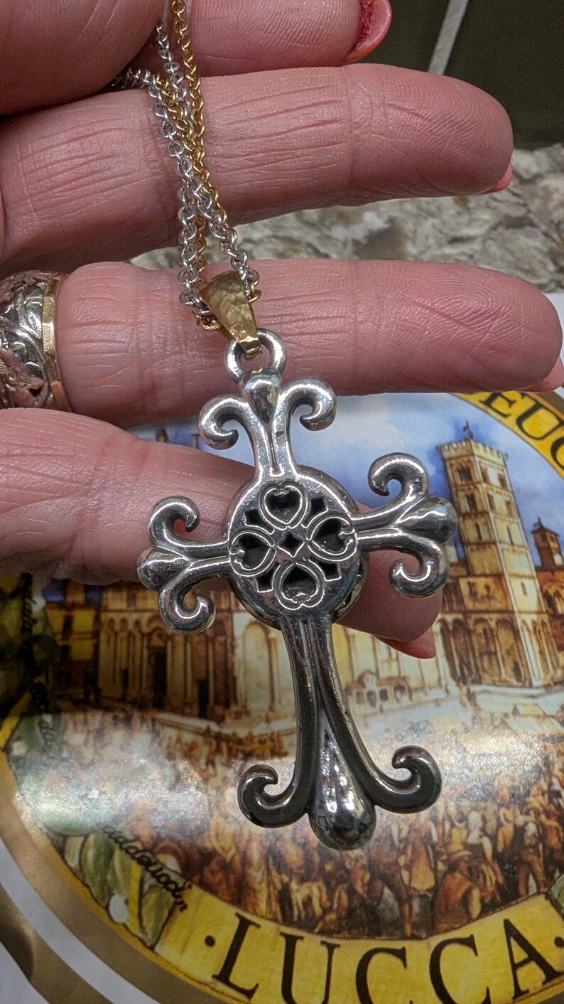 May include: Silver-coloured cross pendant necklace with a decorative design. The cross has a detailed, openwork centre with heart-shaped cutouts and ornate, curved arms. The chain is a mix of silver and gold tones.