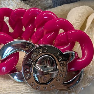 May include: A bright pink plastic chain bracelet with a silver Marc Jacobs logo charm.