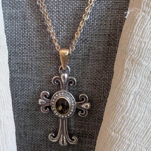May include: Silver-toned cross pendant necklace with a gold-toned bail and a double chain. The cross features an oval, dark-coloured stone surrounded by small clear stones. The cross has ornate, scrolled details.