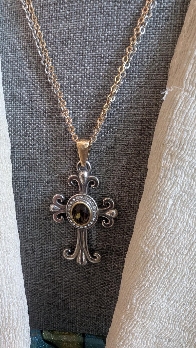 May include: Silver-toned cross pendant necklace with a gold-toned bail and a double chain. The cross features an oval, dark-coloured stone surrounded by small clear stones. The cross has ornate, scrolled details.