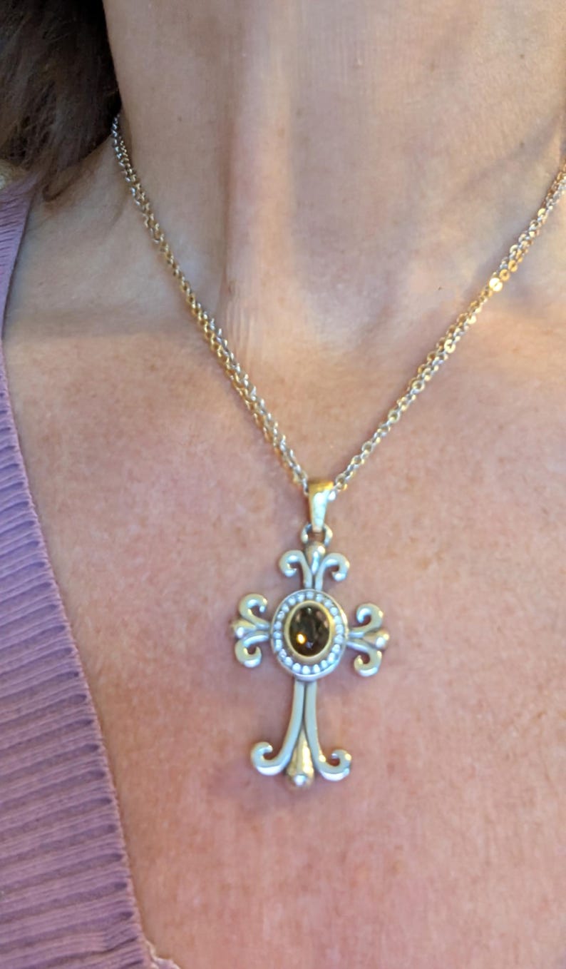 May include: A gold-coloured necklace with a cross pendant. The cross has a central brown gemstone surrounded by small white stones. The cross design has ornate, swirling details. The necklace is worn against a person's skin and a purple top.