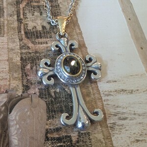 May include: Silver-tone cross pendant necklace with a gold-tone bail. The cross features an oval, dark-coloured stone surrounded by small, clear stones. The chain is a mix of silver and gold tones.