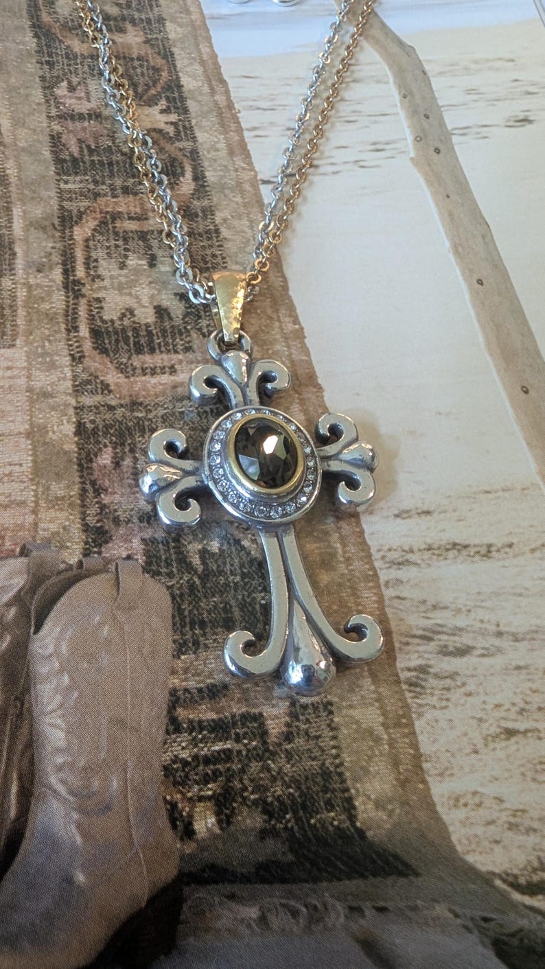 May include: Silver-tone cross pendant necklace with a gold-tone bail. The cross features an oval, dark-coloured stone surrounded by small, clear stones. The chain is a mix of silver and gold tones.