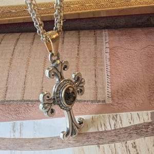 May include: Silver cross pendant necklace with a decorative design and a dark-coloured centre stone. The pendant hangs from a silver chain with a gold-coloured clasp. The cross features ornate detailing against a textured background.
