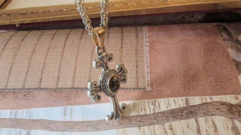 May include: Silver cross pendant necklace with a decorative design and a dark-coloured centre stone. The pendant hangs from a silver chain with a gold-coloured clasp. The cross features ornate detailing against a textured background.