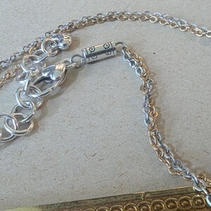 May include: Close-up of a silver-toned necklace with a double chain design. The necklace features a lobster clasp and an extender chain. The chain is connected to a decorative rectangular pendant with a gold-coloured border.