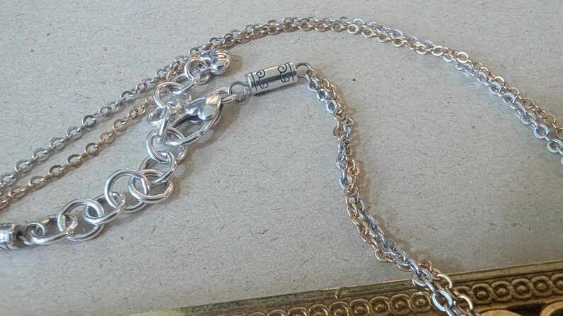 May include: Close-up of a silver-toned necklace with a double chain design. The necklace features a lobster clasp and an extender chain. The chain is connected to a decorative rectangular pendant with a gold-coloured border.