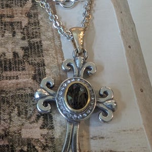 May include: Silver-tone cross pendant necklace with a dark oval stone centre, surrounded by small clear stones. The cross has ornate, curved arms and is suspended from a layered chain.