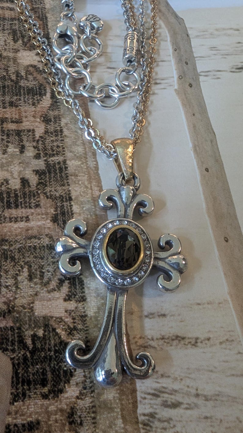 May include: Silver-tone cross pendant necklace with a dark oval stone centre, surrounded by small clear stones. The cross has ornate, curved arms and is suspended from a layered chain.