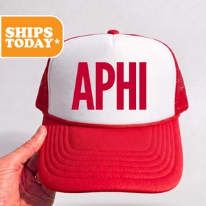 May include: A red and white trucker hat featuring a white front panel with the red text APHI printed in a bold sans-serif font. The hat includes a red brim, a red mesh back, and a red cord detail across the base of the front panel.