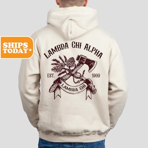 May include: A cream-colored hooded sweatshirt featuring a dark brown graphic on the back. The design includes an axe, pine cones, and branches with the text LAMBDA CHI ALPHA, EST. 1909, and a banner reading LAMBDA CHI across the center of the graphic.