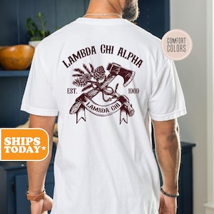 May include: A white short-sleeve t-shirt featuring a dark red graphic on the back. The design includes an axe, pine branches, and pinecones with the text LAMBDA CHI ALPHA, EST. 1909, and a banner reading LAMBDA CHI. The shirt has a Comfort Colors label.