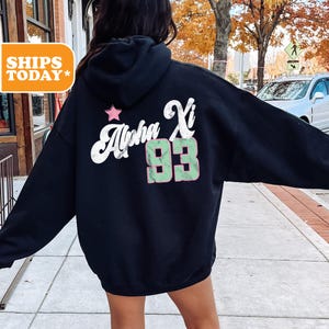 May include: Black hooded sweatshirt with the text "Alpha Xi 93" in white and green. A pink star is above the text. The sweatshirt is worn by a person standing outside on a sidewalk.
