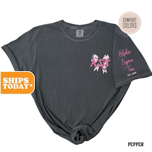 May include: A dark gray t-shirt with a pink bow and the letters "ΑΣΤ" in the center. The text "Alpha Sigma Tau Est. 1899" is on the right sleeve.