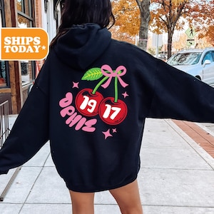 May include: Black hoodie with a pink bow and two red cherries with the numbers 19 and 17 on them. The word "Sophie" is written in pink below the cherries.