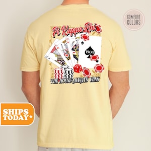 May include: A yellow t-shirt with a graphic of playing cards, dice, and chips. The text "Pi Kappa Phi" and "The House Always Wins" are on the shirt.