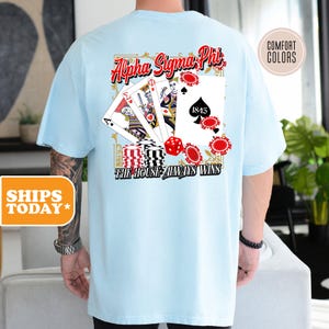 May include: A light blue t-shirt with a red, white, and black design featuring playing cards, dice, and chips. The text "Alpha Sigma Phi" and "The House Always Wins" is printed on the shirt.