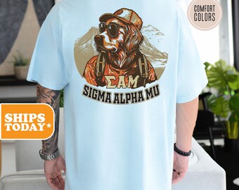 May include: Light blue t-shirt featuring a graphic of a dog wearing sunglasses and a hat, with the text "ΣAM SIGMA ALPHA MU". The dog is set against a mountain backdrop. The shirt is a comfort color.