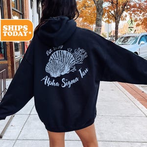 May include: Black hooded sweatshirt with a white graphic of seashells and coral. The text "Est. 1899 Alpha Sigma Tau" is printed on the back.