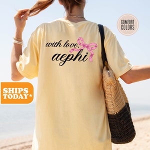 May include: A yellow t-shirt with the text "with love, aephi" and a pink bow on the back.