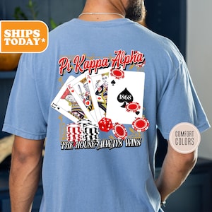 May include: A light blue t-shirt with a graphic of playing cards and the text "Pi Kappa Alpha The House Always Wins".