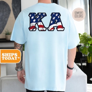 May include: Light blue t-shirt with the letters "KA" in white and red, styled to look like the American flag.