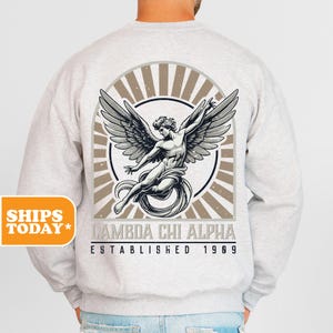 May include: A light gray crewneck sweatshirt featuring a large back graphic of a winged figure in a circular frame with sunburst rays. Below the graphic, the text reads LAMBDA CHI ALPHA ESTABLISHED 1909 in a bold, dark, sans-serif font.