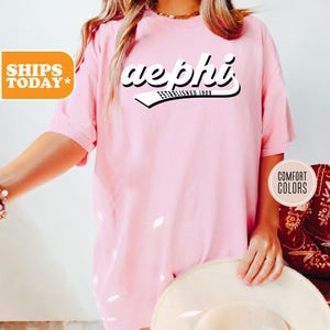 May include: Pink t-shirt with a white graphic that says "aephi established 1909" in a retro font.