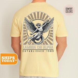 May include: A pale yellow short-sleeve t-shirt featuring a graphic on the back. The design shows a winged figure within a circular sunburst frame, with the text LAMBDA CHI ALPHA and ESTABLISHED 1909 printed in a dark font below the illustration.