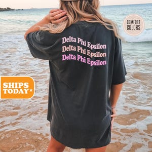 May include: A charcoal grey short-sleeve t-shirt featuring the text Delta Phi Epsilon printed in three stacked rows on the back. The text appears in alternating shades of light pink and peach, with a wavy, curved font style across the upper back.