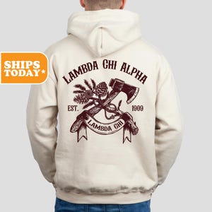 May include: A cream-colored hooded sweatshirt with a maroon graphic of crossed axes and pine branches. The graphic includes the text "Lambda Chi Alpha", "Est. 1909", and "Lambda Chi".