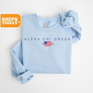 May include: A light blue sweatshirt with the text "ALPHA CHI OMEGA" above a small American flag and the numbers "18" and "85". The sweatshirt has a crew neck and long sleeves.
