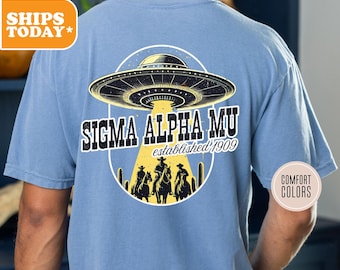 May include: A light blue t-shirt featuring a graphic of a UFO and cowboys riding horses. The text "Sigma Alpha Mu established 1909" is printed on the shirt.