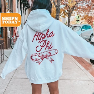 May include: A light blue hooded sweatshirt with the text "Alpha Phi" in red cursive lettering and a red safety pin with a bow on the back.