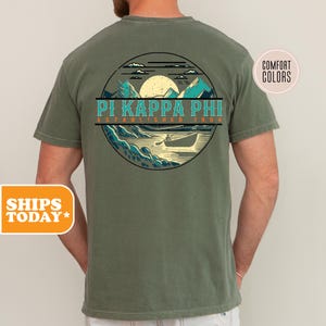 May include: A green short-sleeved t-shirt with a vintage-style graphic of a lake scene with mountains and a canoe. The graphic includes the text "Pi Kappa Phi Established 1904".