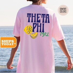 May include: A light pink short-sleeve t-shirt featuring the dark blue text THETA PHI with the green number 1912 and two yellow lemon illustrations on the back. A circular badge in the top right corner contains the text COMFORT COLORS.