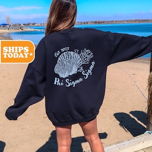 May include: A navy blue sweatshirt with a white graphic of seashells and coral. The text "Est. 1973 Phi Sigma Sigma" is printed on the back of the sweatshirt.