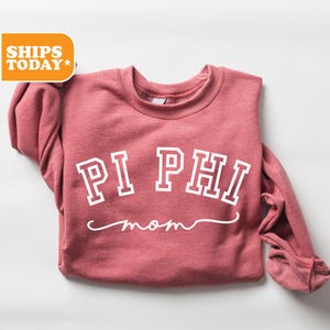 May include: A maroon sweatshirt with the words "PI PHI mom" in white block letters and script. The sweatshirt has a crew neck and long sleeves. The text is centered on the front of the garment.