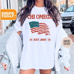 May include: White t-shirt with a red, white, and blue American flag graphic and the text "CHI OMEGA EST. 1895".