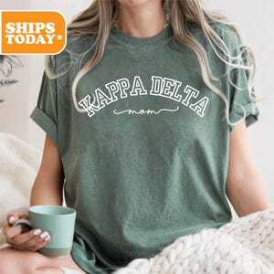 May include: A sage green t-shirt with the text "KAPPA DELTA mom" in white. The shirt has rolled-up sleeves and is made of a soft material. A light blue mug is held in the hand.