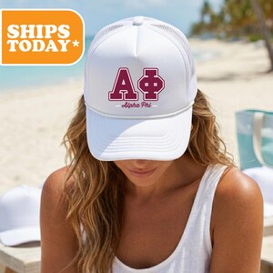 May include: A white trucker hat featuring a mesh back and a solid front panel. The front displays the Greek letters Alpha Phi in a maroon collegiate font with a white outline, accompanied by the text Alpha Phi printed below in a smaller font.