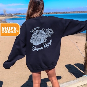 May include: A navy blue sweatshirt with a white graphic of seashells and coral. The text "Est 1874" and "Sigma Kappa" are also printed on the sweatshirt.