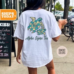 May include: White t-shirt with a turtle design in teal, yellow, and blue. The text "Alpha Sigma Tau" is printed below the turtles. The shirt is a Comfort Colors brand.