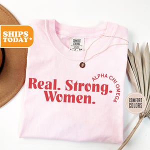 May include: Pink t-shirt with the text "Real. Strong. Women." in red. The text "ALPHA CHI OMEGA" is in a circle around the words "Strong. Women."