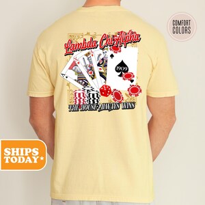 May include: A pale yellow t-shirt featuring a back graphic of playing cards, red poker chips, and a red die. The text reads Lambda Chi Alpha and The House Always Wins. The design includes a spade symbol with the number 1909 inside the card.